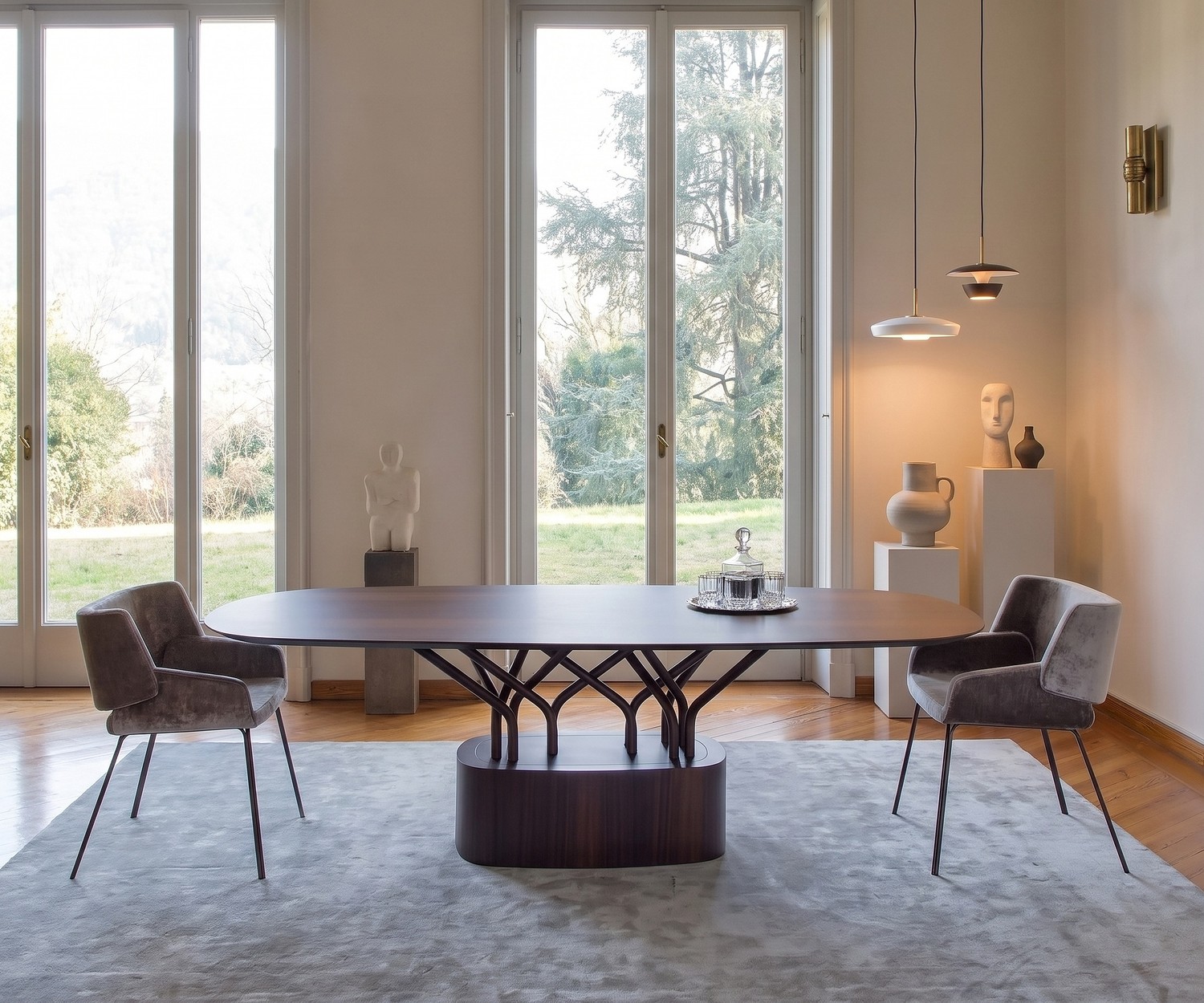 al2 wood-oo Design Esstisch oval Ovaler al2 wood-oo Esstisch aus Holz im modernen Esszimmer mit zwei grauen Samtsesseln vor großen Fenstern.