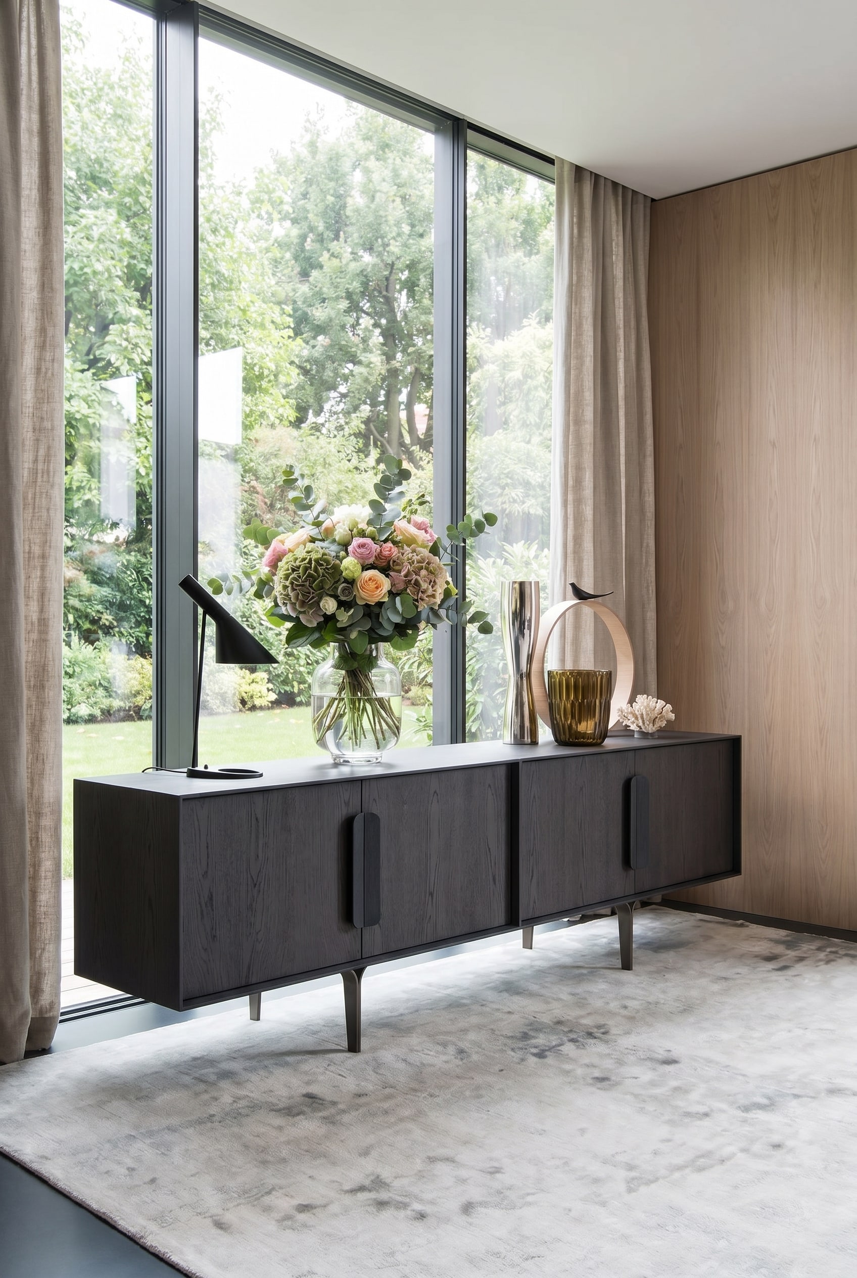 Buffet design fonc&eacute; devant de grandes fen&ecirc;tres, sur lequel repose un vase statement lumineux et clair avec des fleurs printani&egrave;res