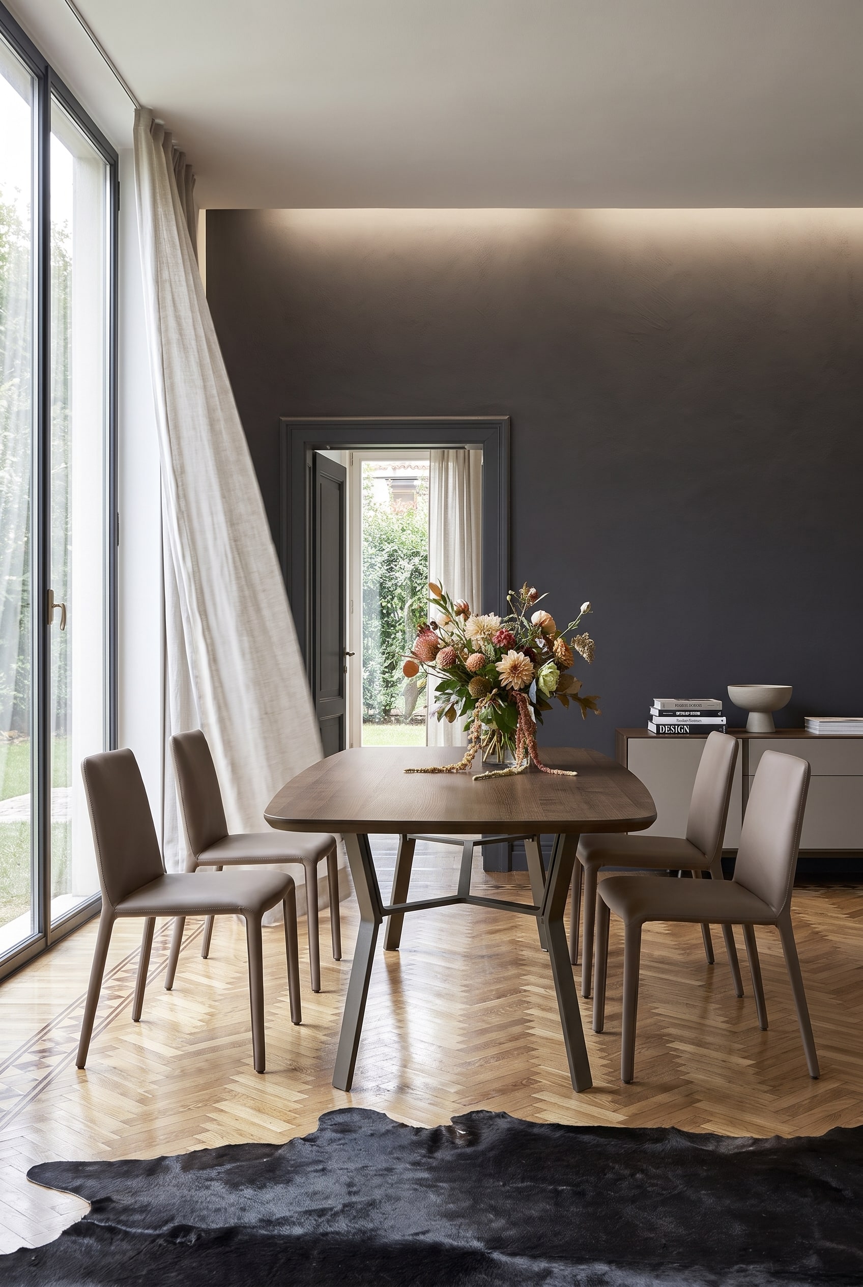 Salle &agrave; manger minimaliste avec mur d'accent sombre, table en bois sur pi&egrave;tement en fer et quatre chaises en cuir couleur taupe