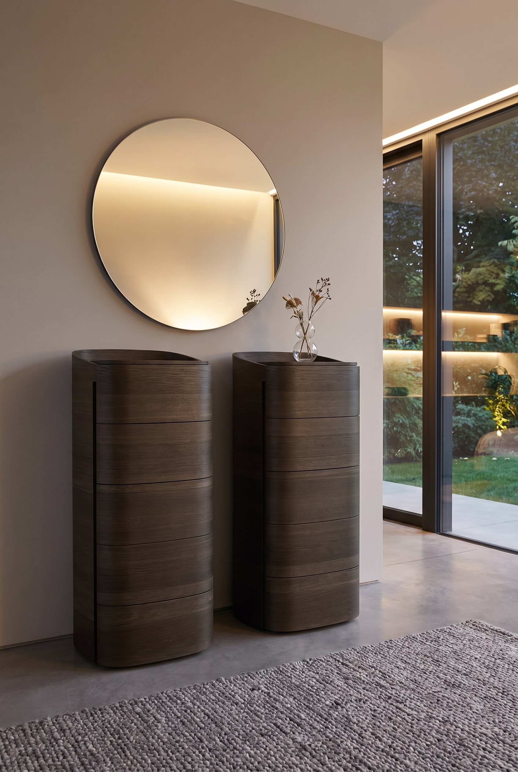 Commode haute et arrondie en bois fonc&eacute;, surmont&eacute;e d'un grand miroir rond minimaliste au mur, avec un petit vase devant