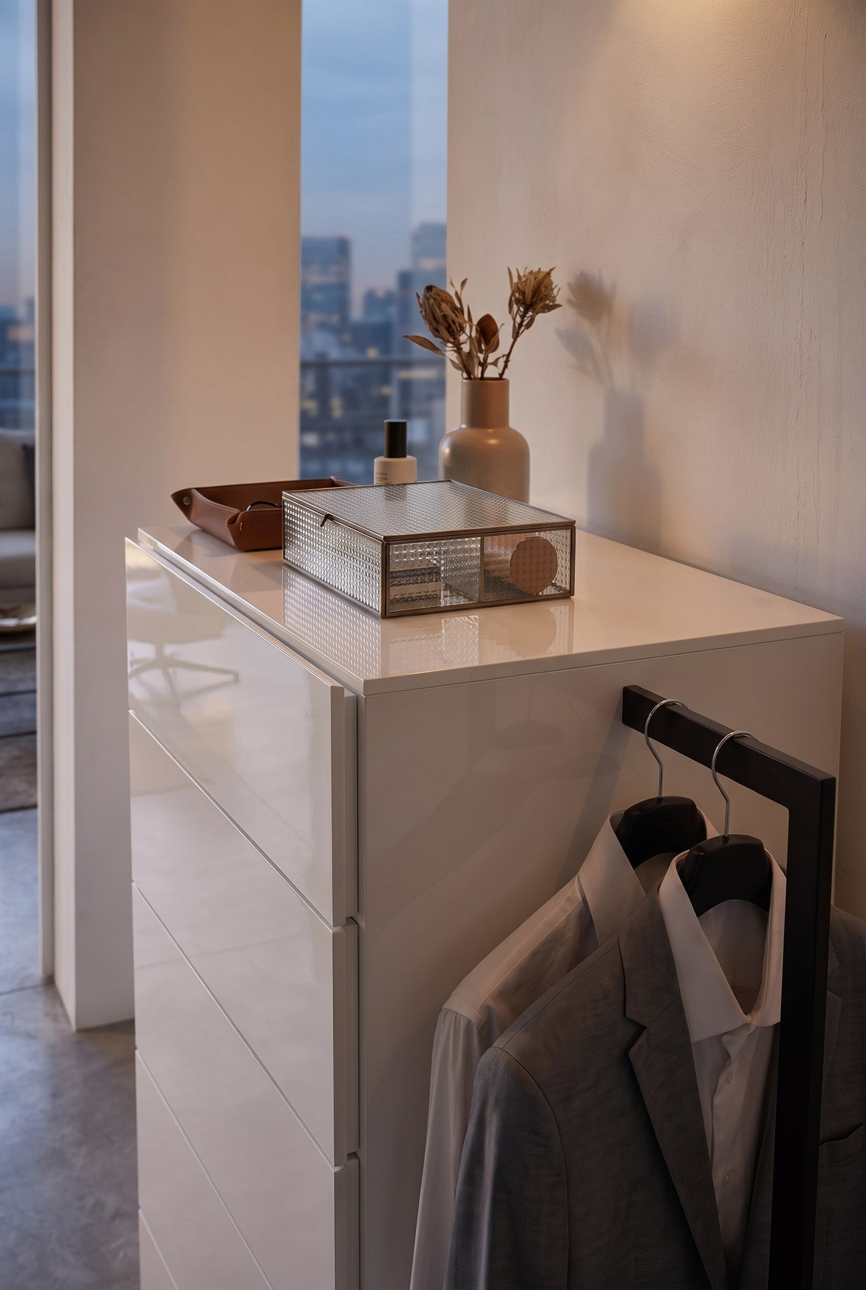 Vue d&eacute;taill&eacute;e d'une commode blanche avec une bo&icirc;te en verre cannel&eacute;, un plateau en cuir et une tringle &agrave; v&ecirc;tements lat&eacute;rale avec une veste grise.