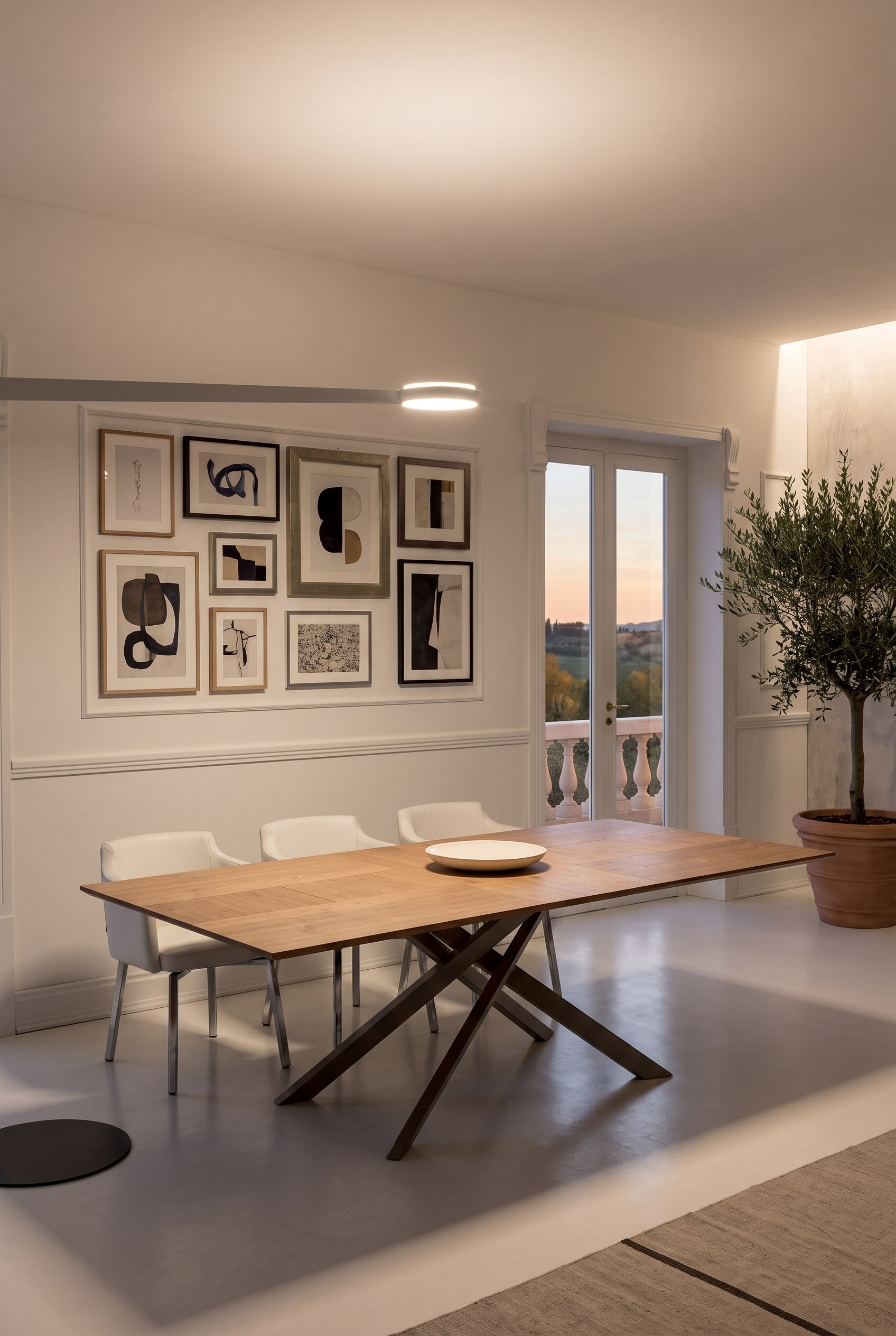 Grande table en bois extensible avec pi&egrave;tement m&eacute;tallique crois&eacute; dans une salle &agrave; manger lumineuse, encadr&eacute;e par une galerie de tableaux parfaitement align&eacute;e
