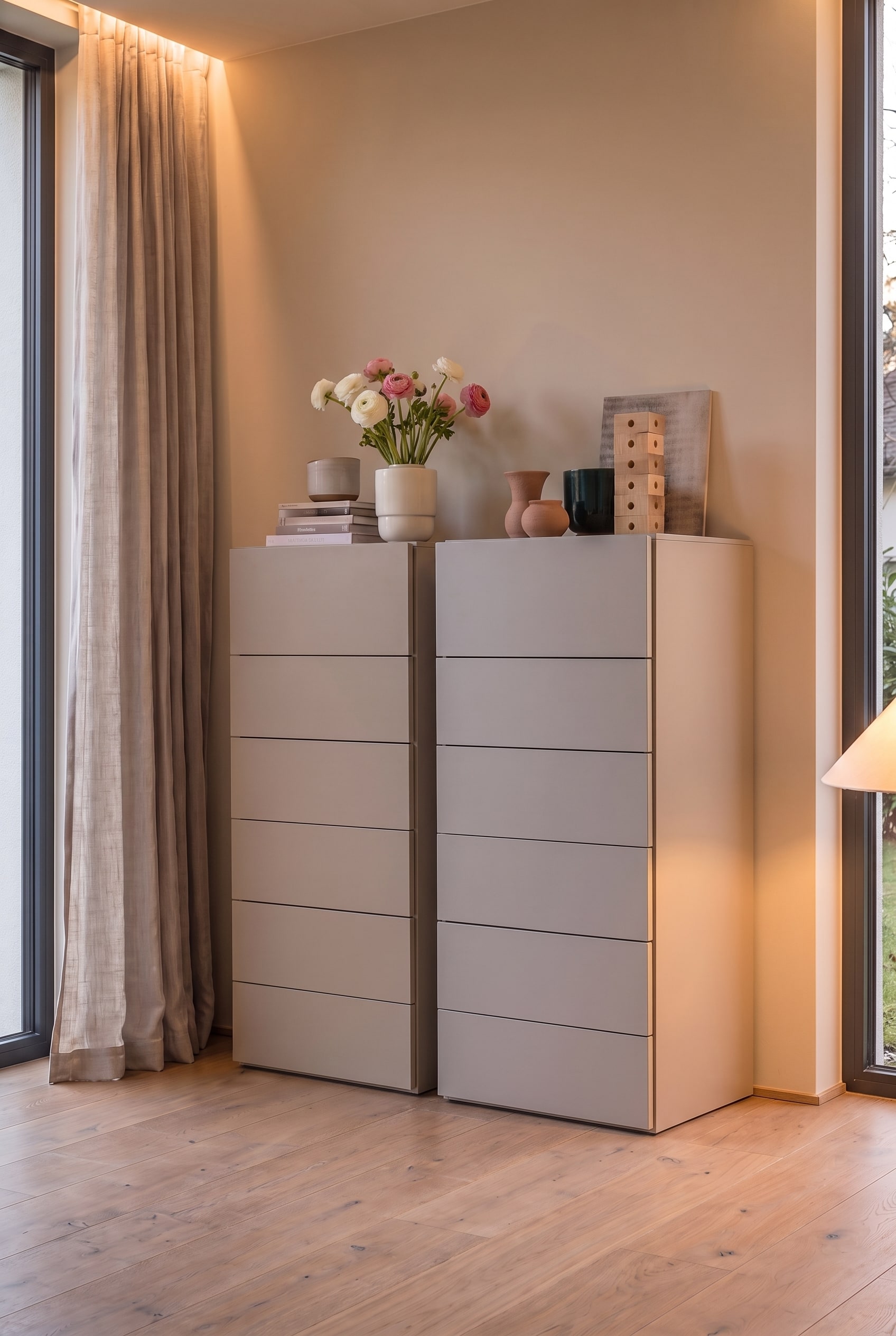 Zwei identische greige Hochkommoden nebeneinander im Schlafzimmer, beleuchtet von warmem Umgebungslicht.