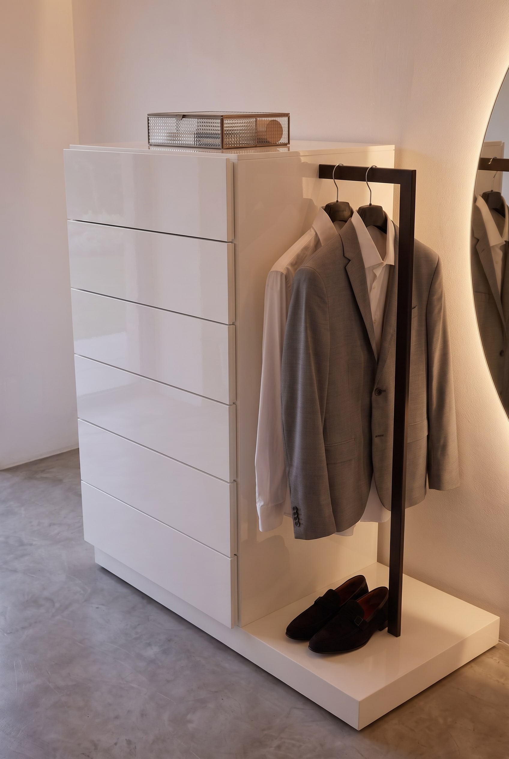 Commode blanche avec tringle de penderie noire latérale. Une veste grise est suspendue au-dessus d'une paire de chaussures foncées sur la plinthe.