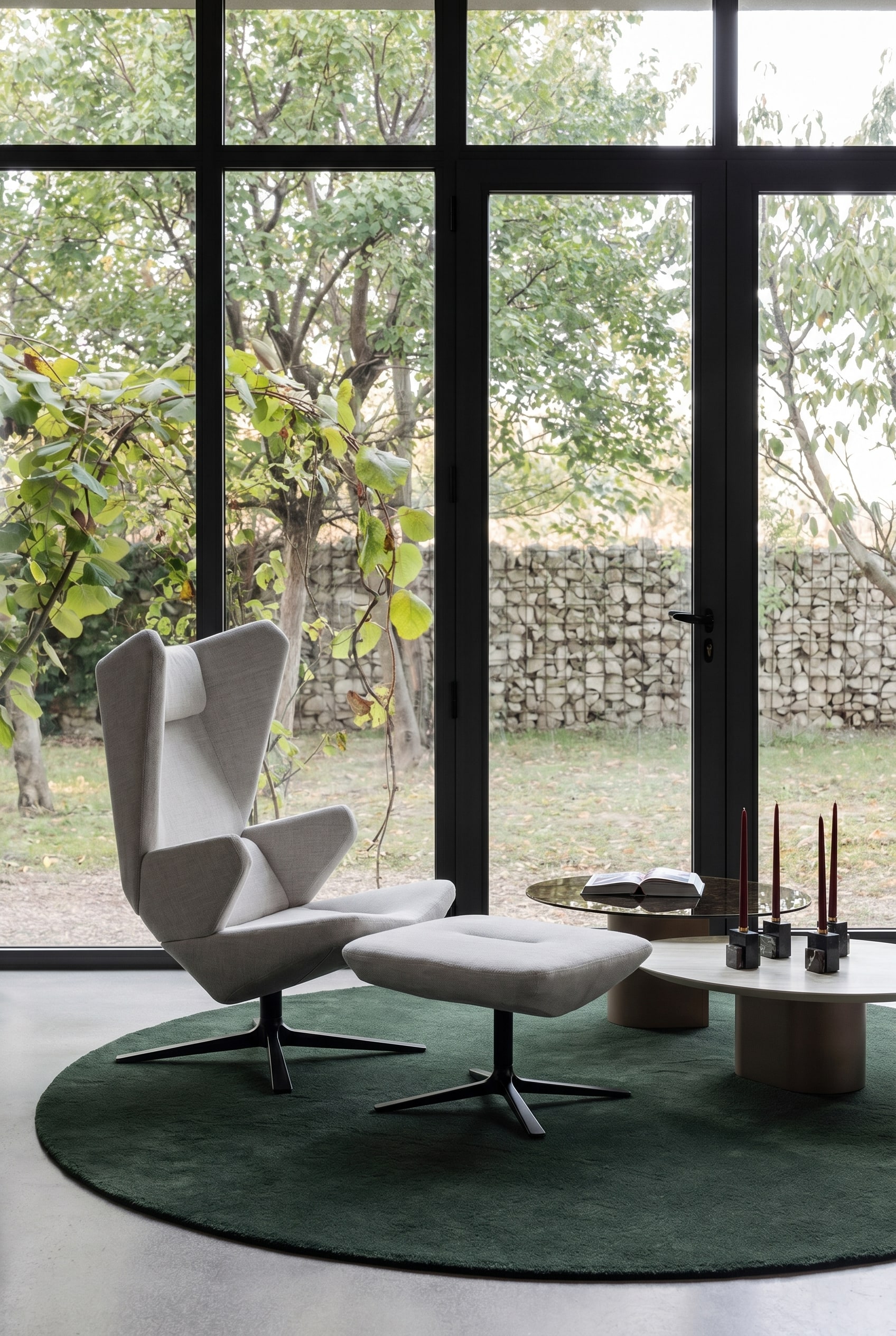 Fauteuil sculptural gris &agrave; dossier haut sur un tapis rond vert fonc&eacute; dans un salon minimaliste avec de grandes fen&ecirc;tres