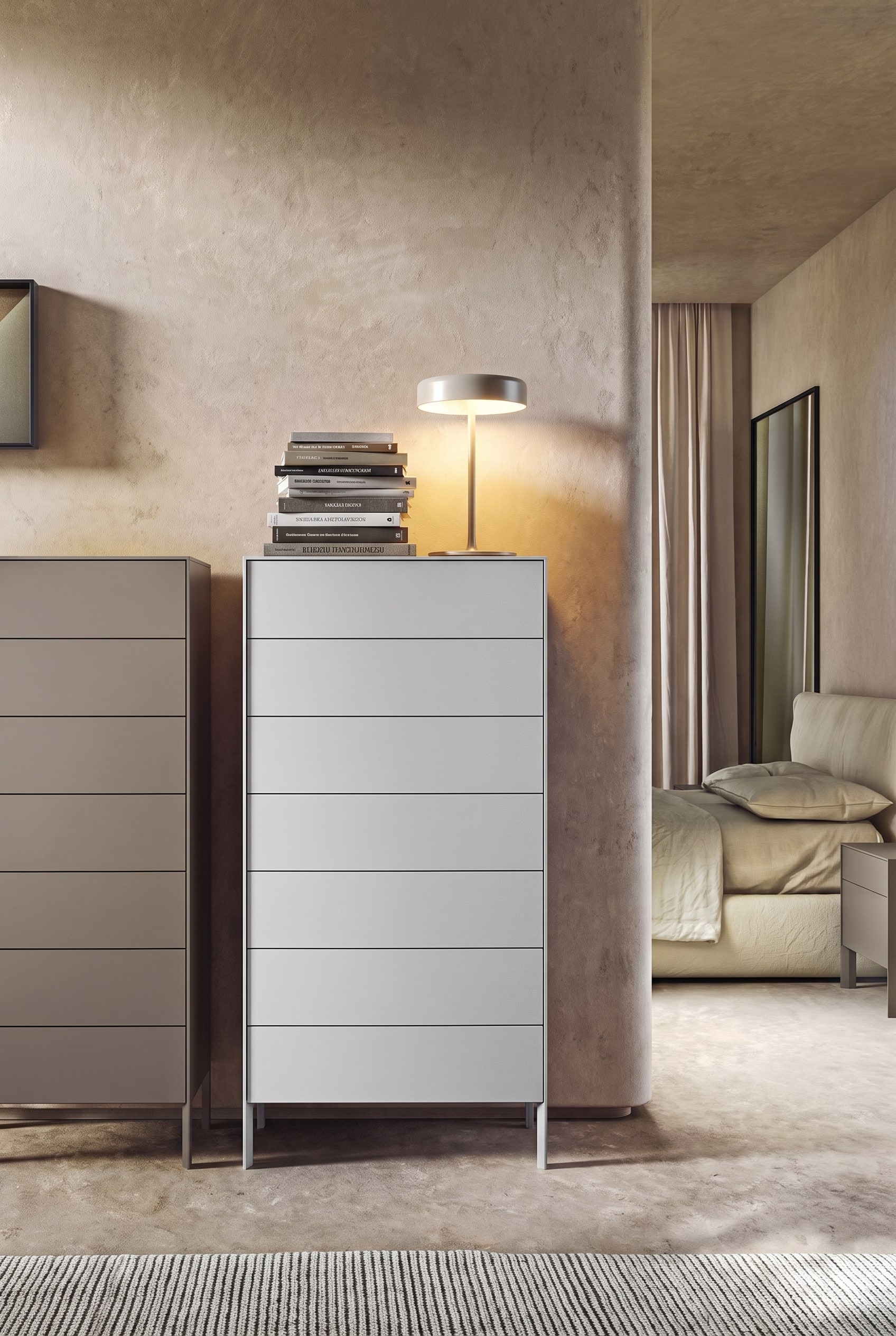 Chambre minimaliste avec une commode grise haute, sur le c&ocirc;t&eacute; gauche une pile massive de livres et une lampe de table, le c&ocirc;t&eacute; droit vide